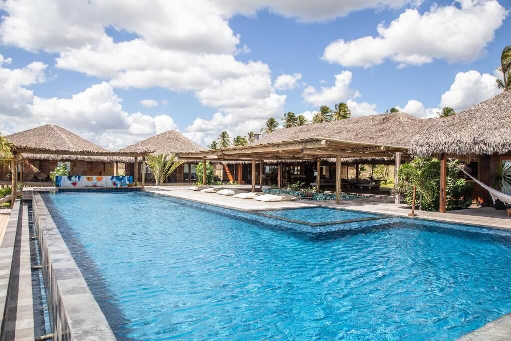Una piscina con estructuras de techo de paja y palmeras al fondo.