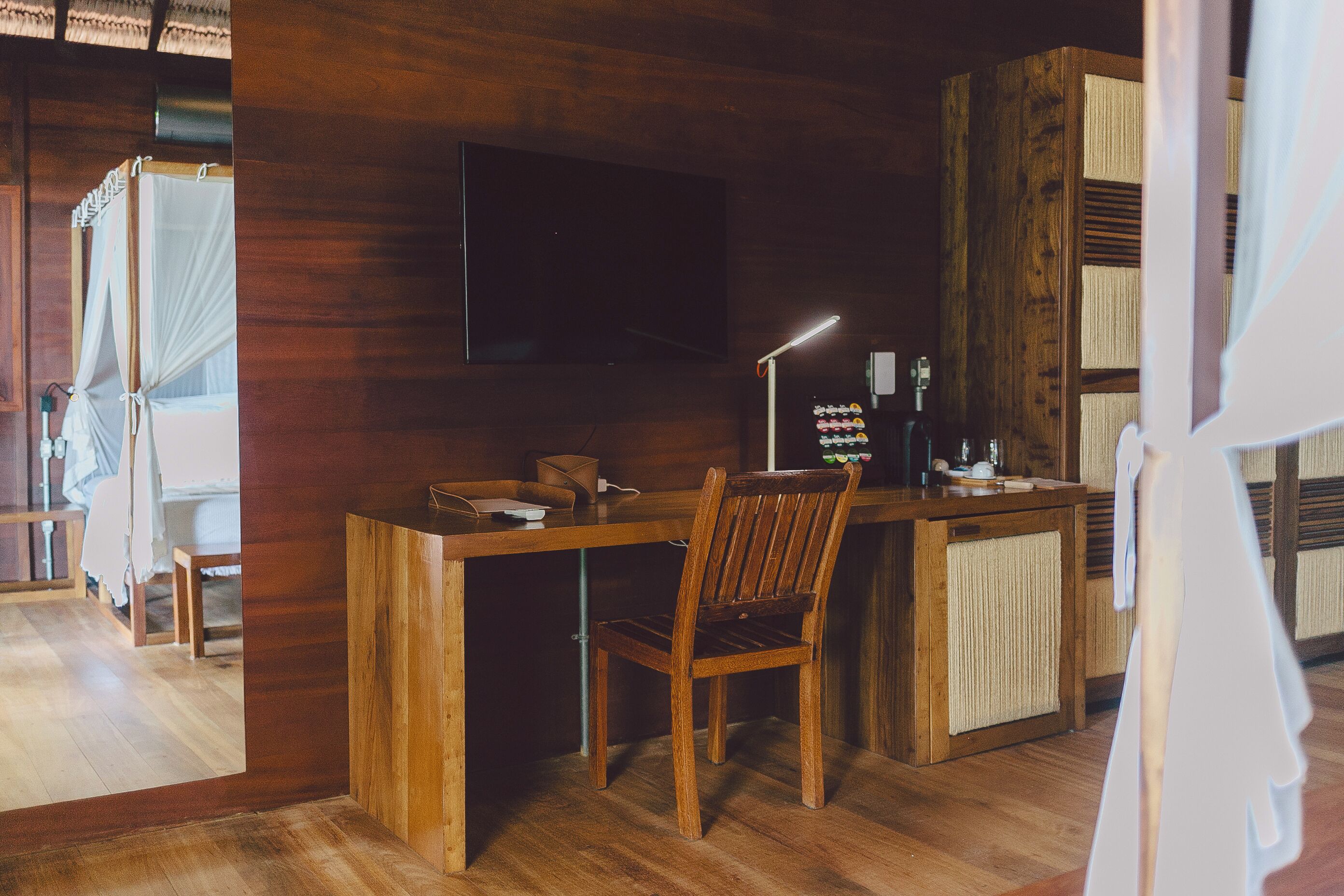 Un bureau en bois avec une chaise, un téléviseur fixé au mur et une table de chevet avec une lampe.