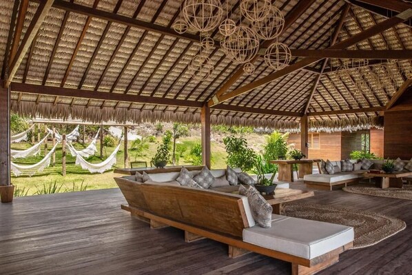 A spacious outdoor lounge area with wooden furniture, a thatched roof, and hanging wicker lamps. - Kabana Hotel (Cruz)