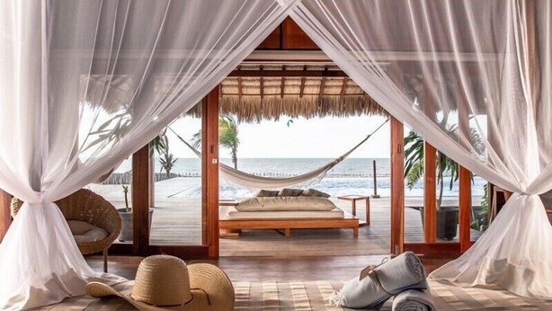 A beachside bedroom with a hammock bed, a straw hat, and a rolled towel on the floor. - Kabana Hotel (Cruz)
