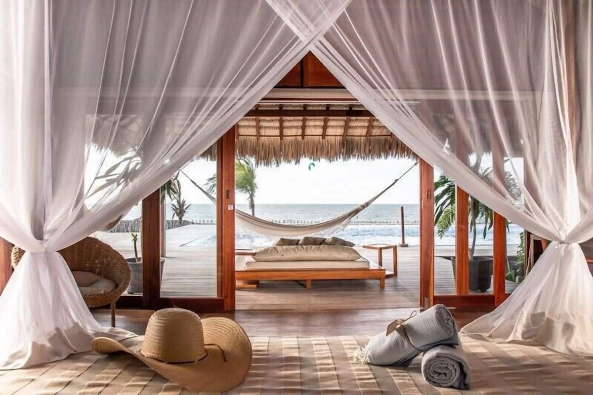 A beachside bedroom with a hammock bed, a straw hat, and a rolled towel on the floor.