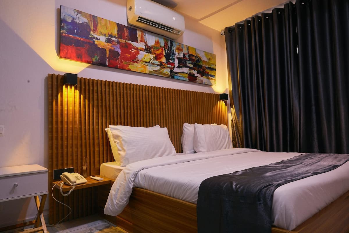 Una habitación de hotel con cabecera de madera, una cama con ropa blanca, una mesita de noche con teléfono y un cuadro sobre la cama.