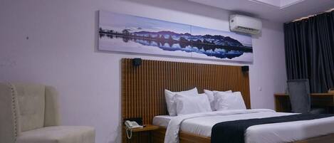 Quarto de hotel com cama, uma cadeira, mesa de cabeceira com telefone, ar condicionado fixado na parede e uma grande pintura de paisagem.
