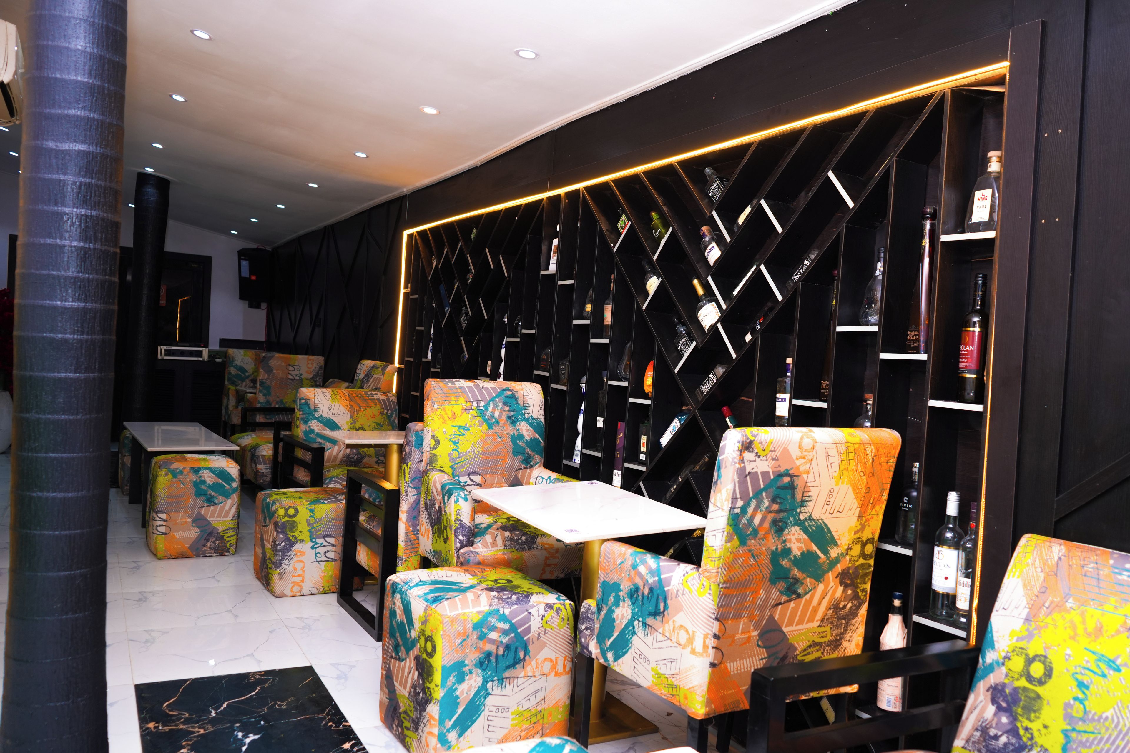 A modern interior with colorful, graffiti-style upholstered chairs and tables, a black wall with a geometric shelving unit, and a ceiling with recessed lighting.