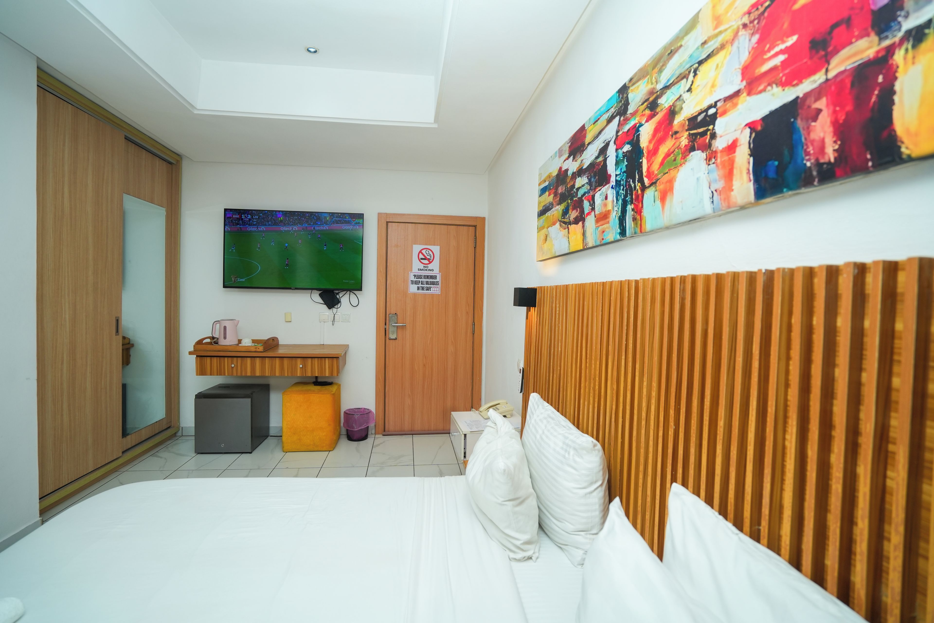 A hotel room with a bed, a TV mounted on the wall, a wooden headboard, a desk with a chair, a trash bin, and a door with a no-smoking sign.