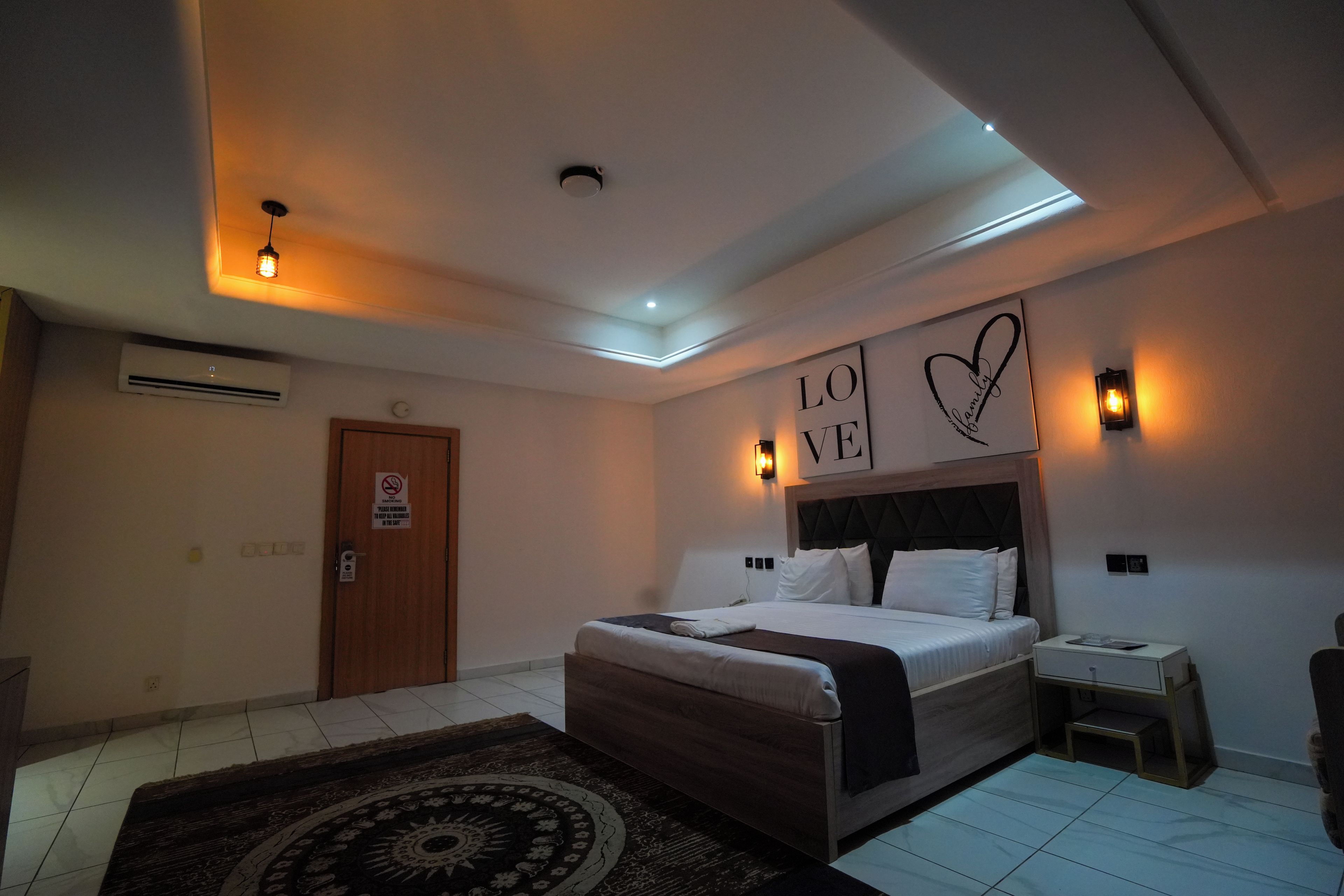 Une chambre d’hôtel comprenant un lit, des tables de chevet, une lampe fixée au mur, une porte avec un panneau « interdiction de fumer » et un luminaire au plafond.