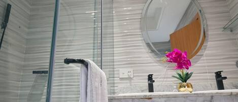 Une salle de bain avec une douche à parois de verre, un comptoir en marbre, un miroir rond et une orchidée en pot.