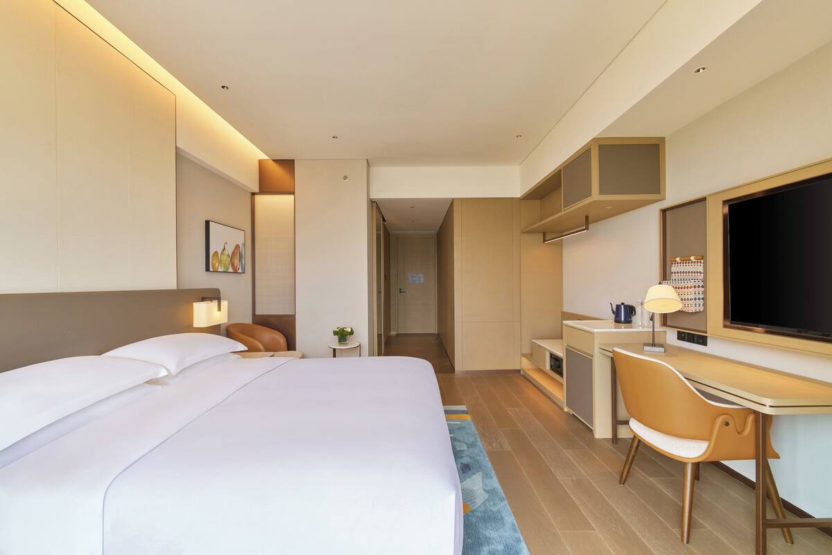 A modern hotel room with a large bed, a desk, a chair, a television, and a small table with a lamp.