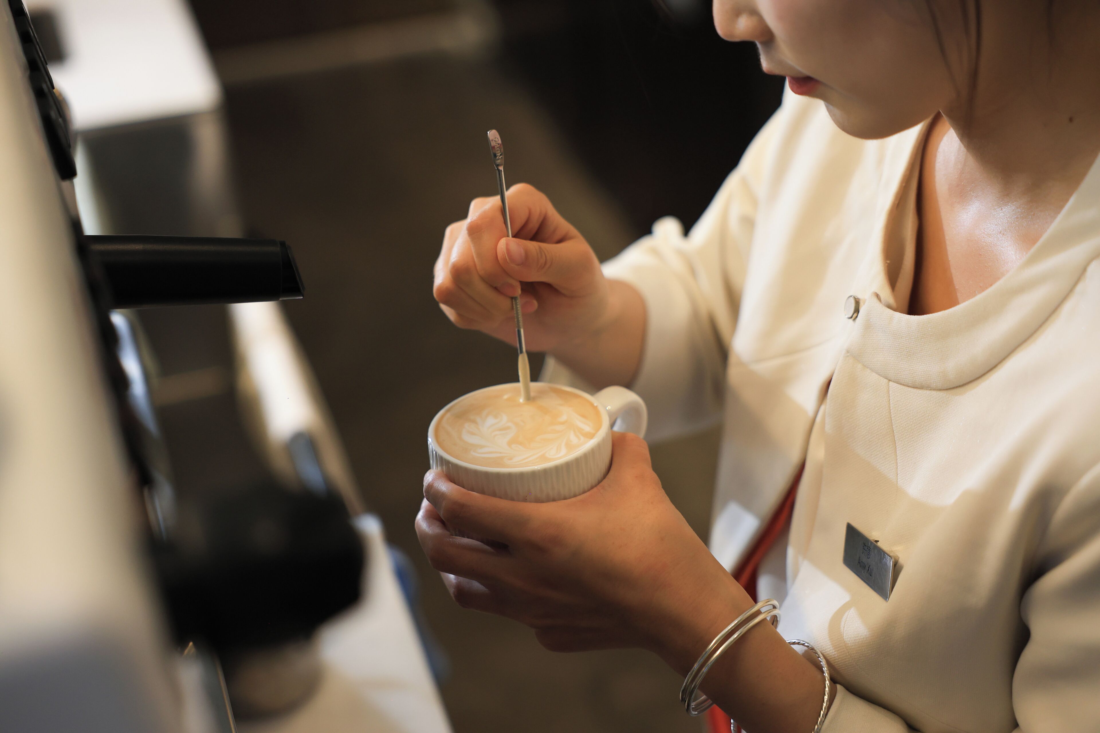 A person holding a cup with a latte art design, wearing a name tag, and a bracelet.