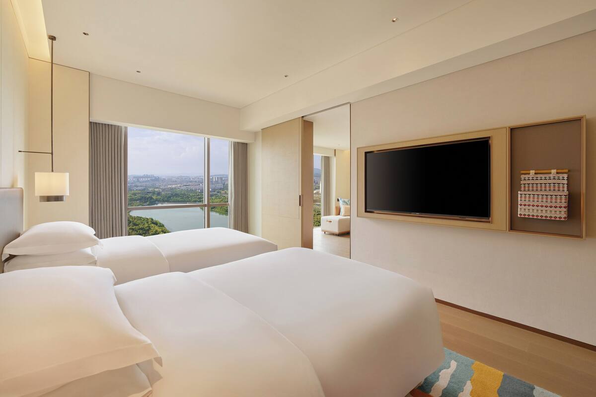 A hotel room with two beds, a TV, a large window with a city view, and a mirror.