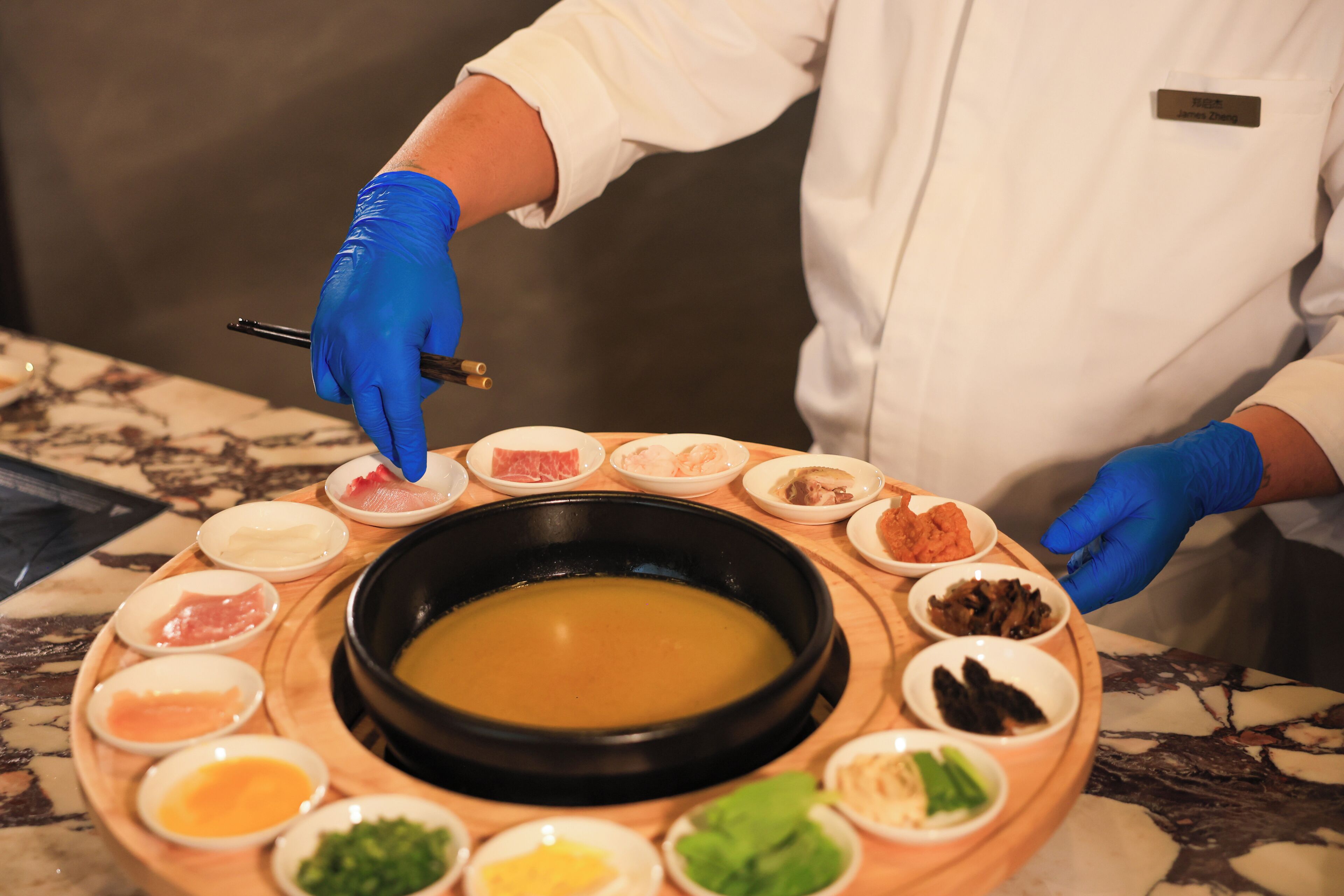 A person in a white uniform with blue gloves serving a hot pot meal surrounded by various condiments on a wooden tray.