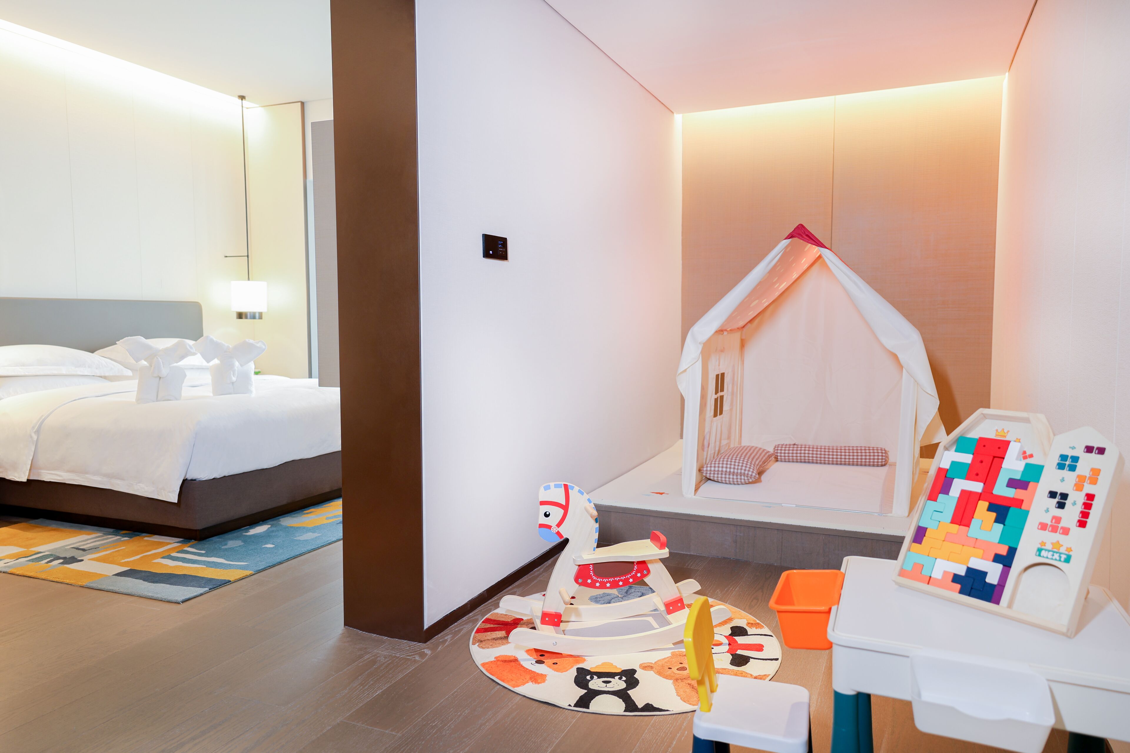 Une chambre d’hôtel avec un lit, un espace de jeux comprenant une chaise suspendue, une table à jeux de puzzle et une petite table avec un jouet.