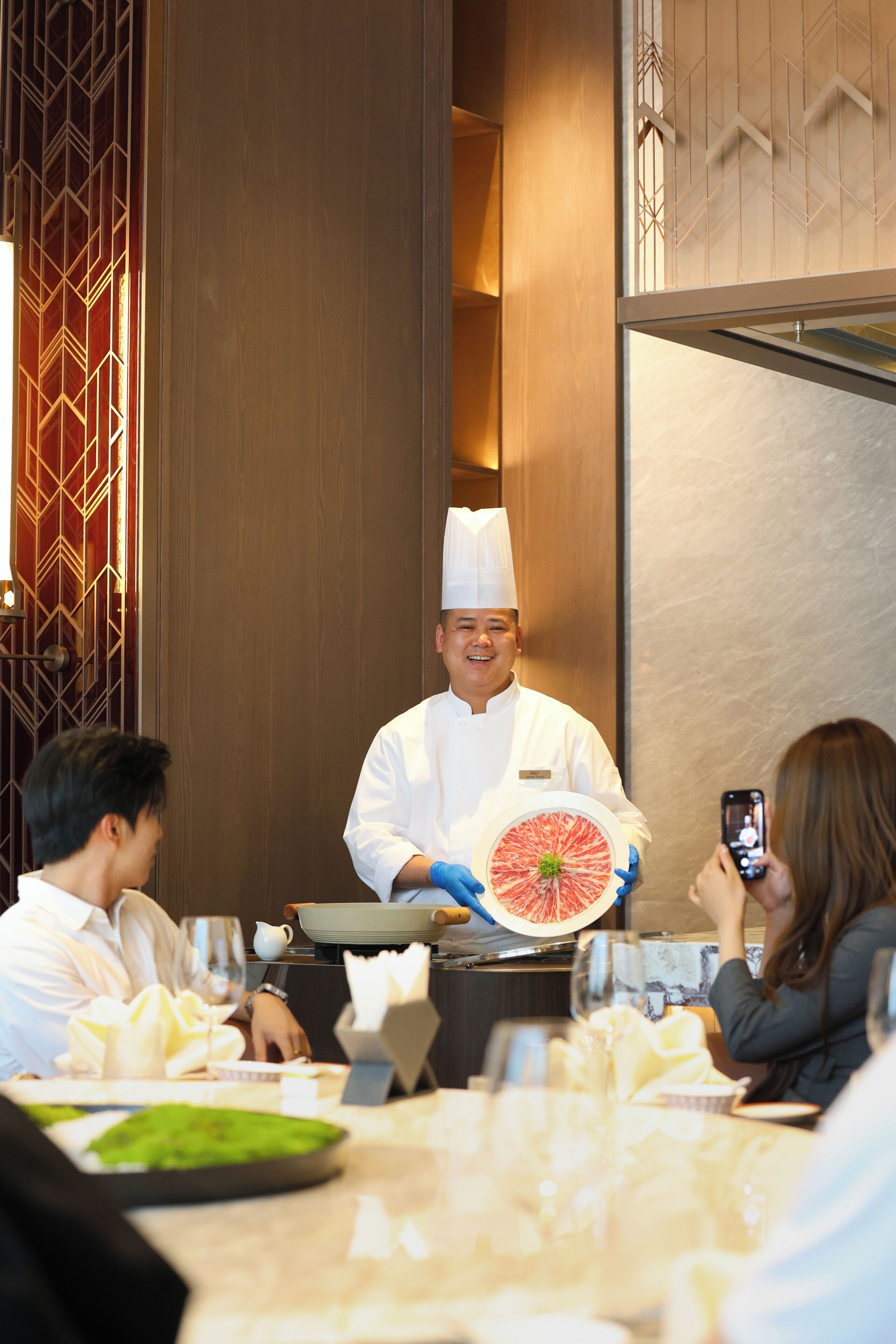 A chef in a white uniform holding a grapefruit, standing in a restaurant with diners.