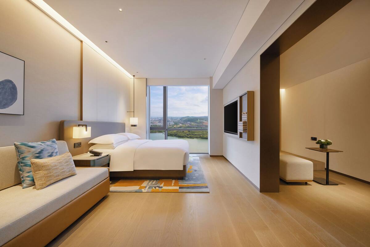 A modern hotel room with a bed, a sofa, a TV, and a view of the city.