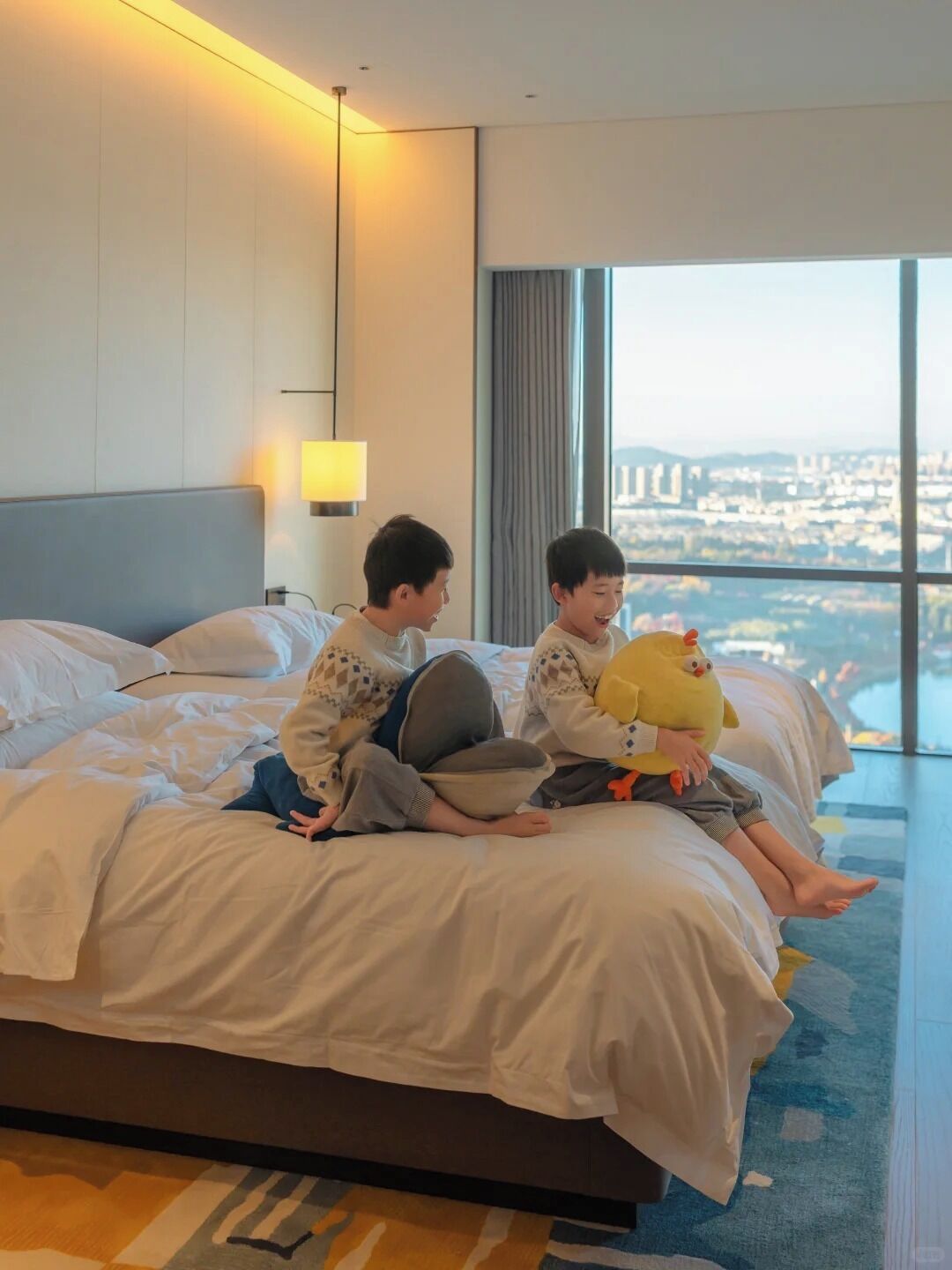 A hotel room with a large bed, a city view, and a lamp on the wall.