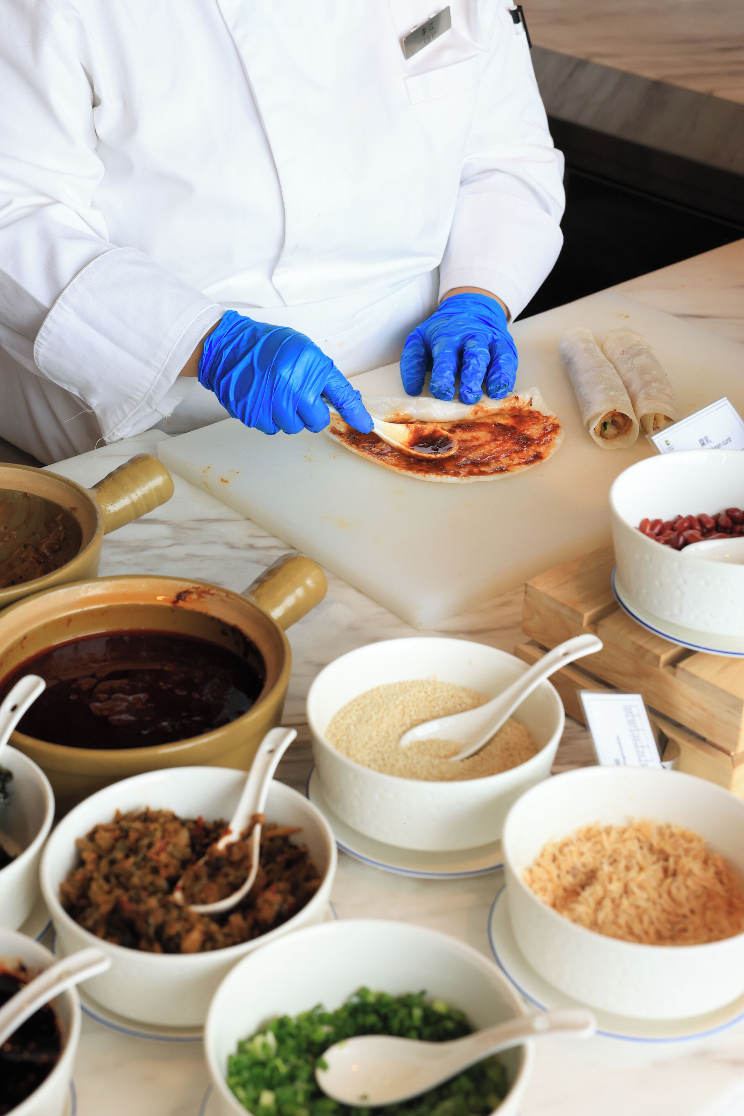 A person in a white coat and blue gloves preparing food on a cutting board with various ingredients in bowls around.
