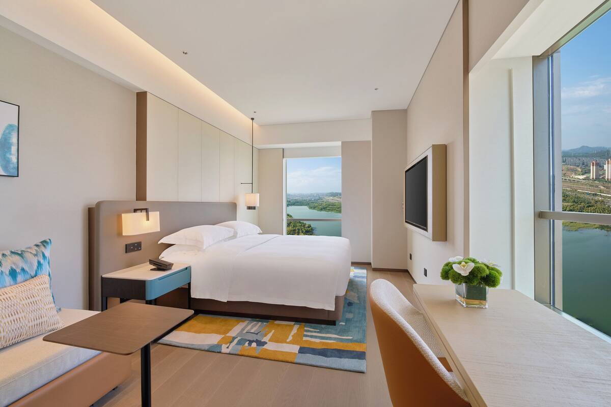 A modern hotel room with a large bed, bedside table, lamp, TV, and a view of a cityscape.