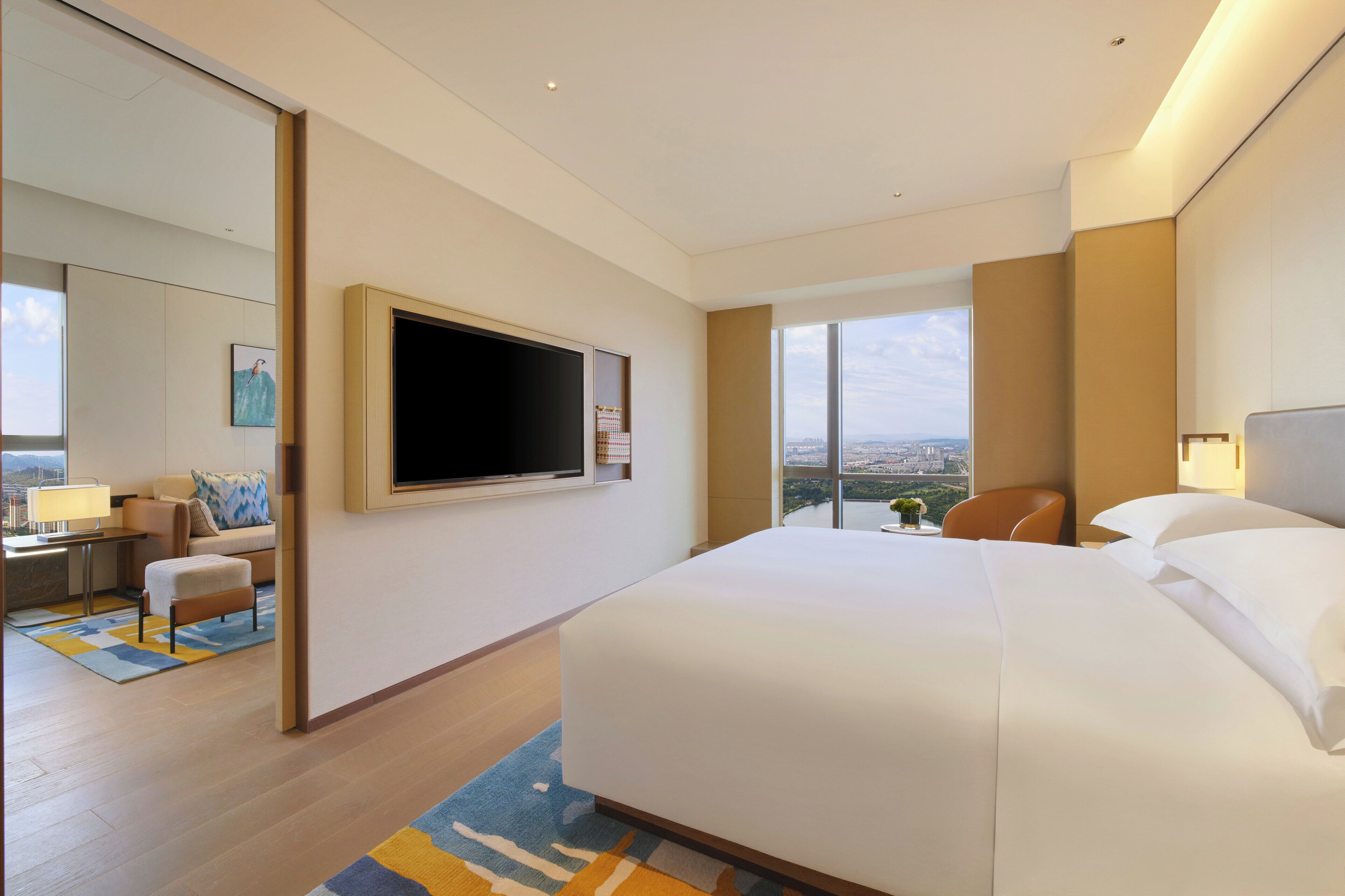 A hotel room with a large bed, a flat-screen TV mounted on the wall, a seating area with a sofa and armchair, and a view of the city through large windows.