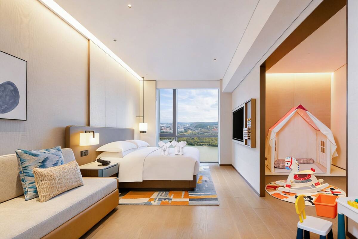 A modern hotel room with a bed, a sofa, a TV, a play area, and a large window offering a city view.