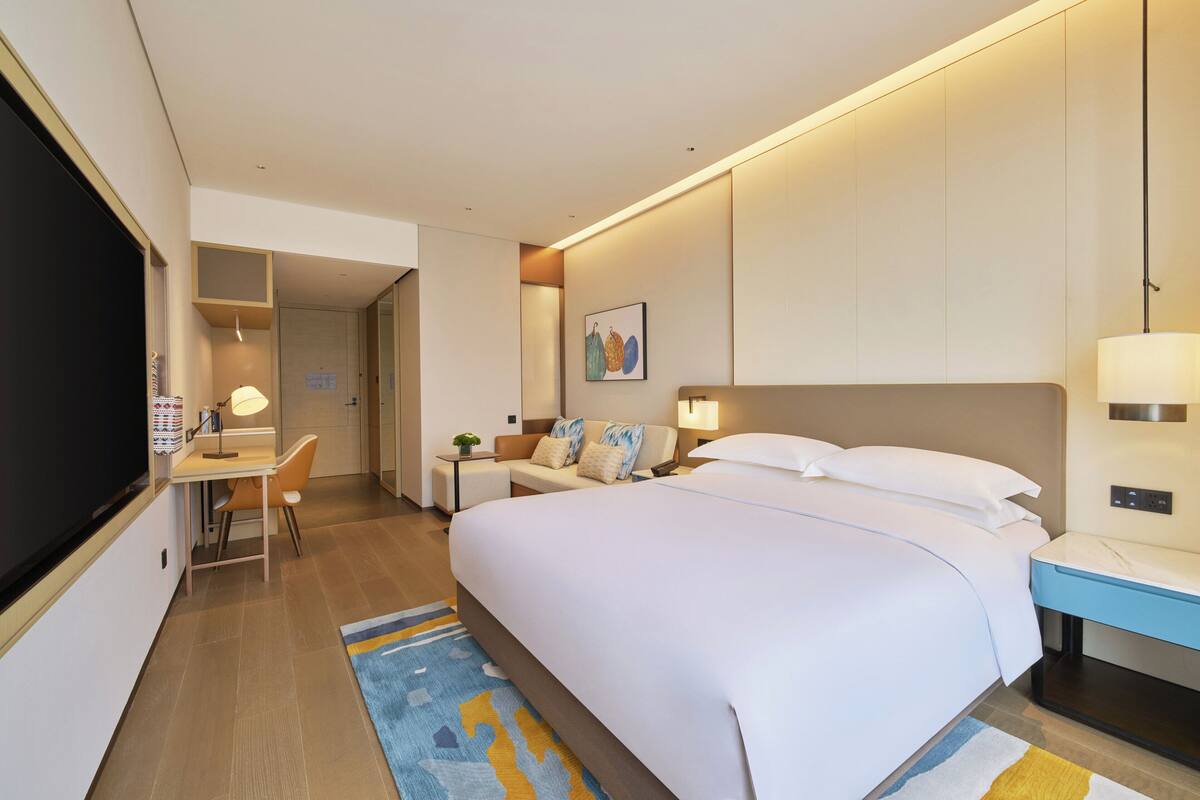 A hotel room with a large bed, a desk with a chair, a TV, and a bedside table with a lamp.