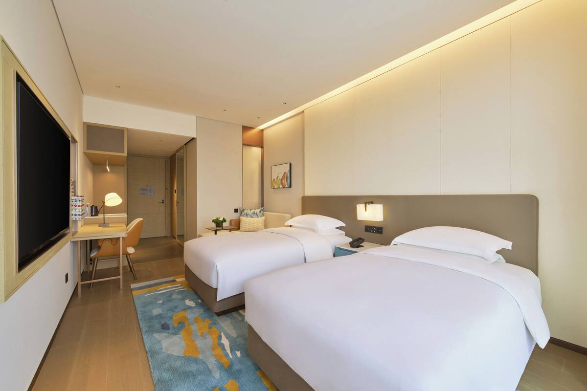 A hotel room with two beds, a desk, a TV, and a small table with a lamp.