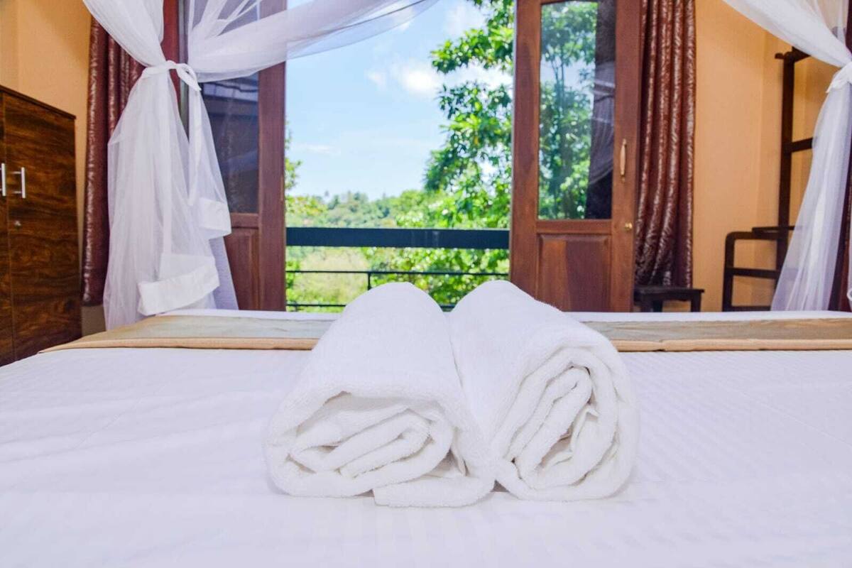 Una cama bien hecha con toallas blancas, un cabecero de madera y vistas al verdor a través de una ventana con cortinas.