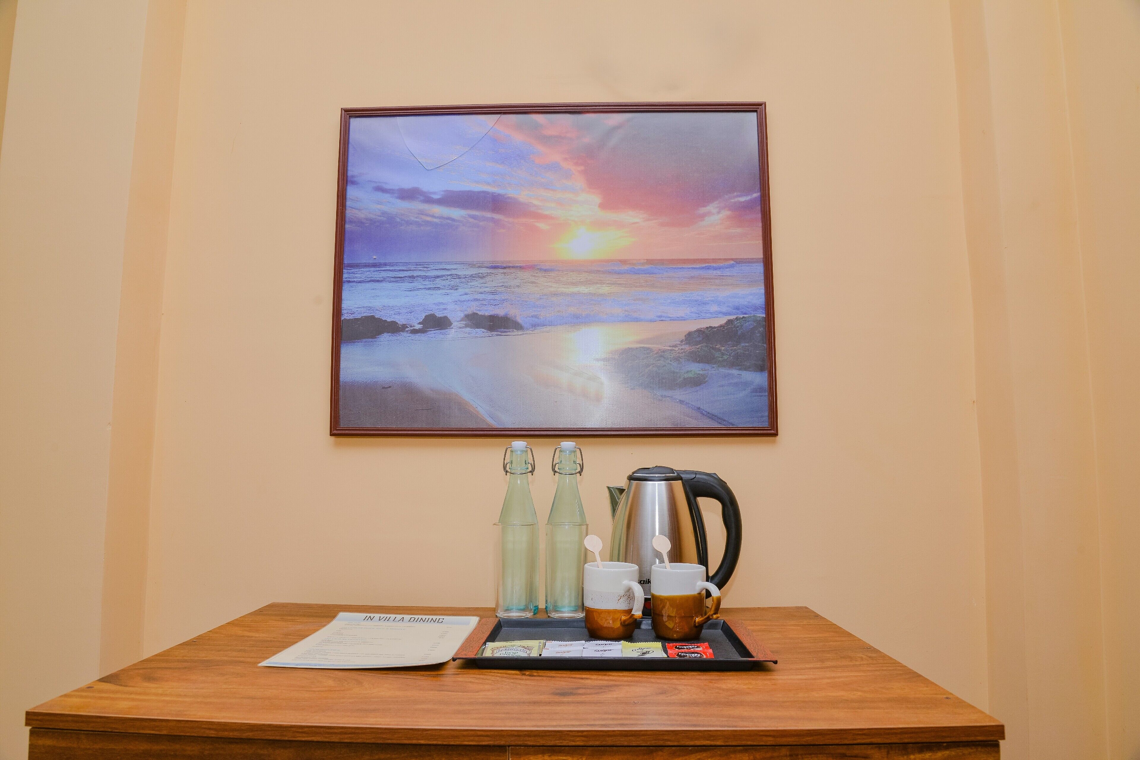 Una mesa de madera con una bandeja que contiene una cafetera, dos tazas y dos botellas, frente a una pared con un cuadro de un atardecer enmarcado.