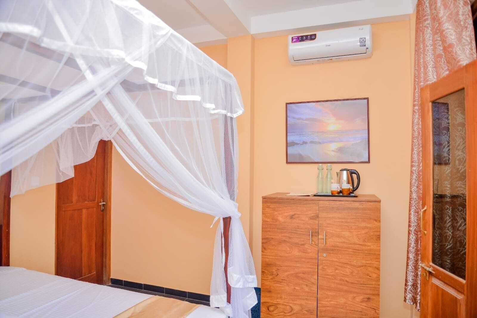 A bedroom with a bed under a mosquito net, a wooden wardrobe, a painting of a sunset, and an air conditioning unit on the wall.