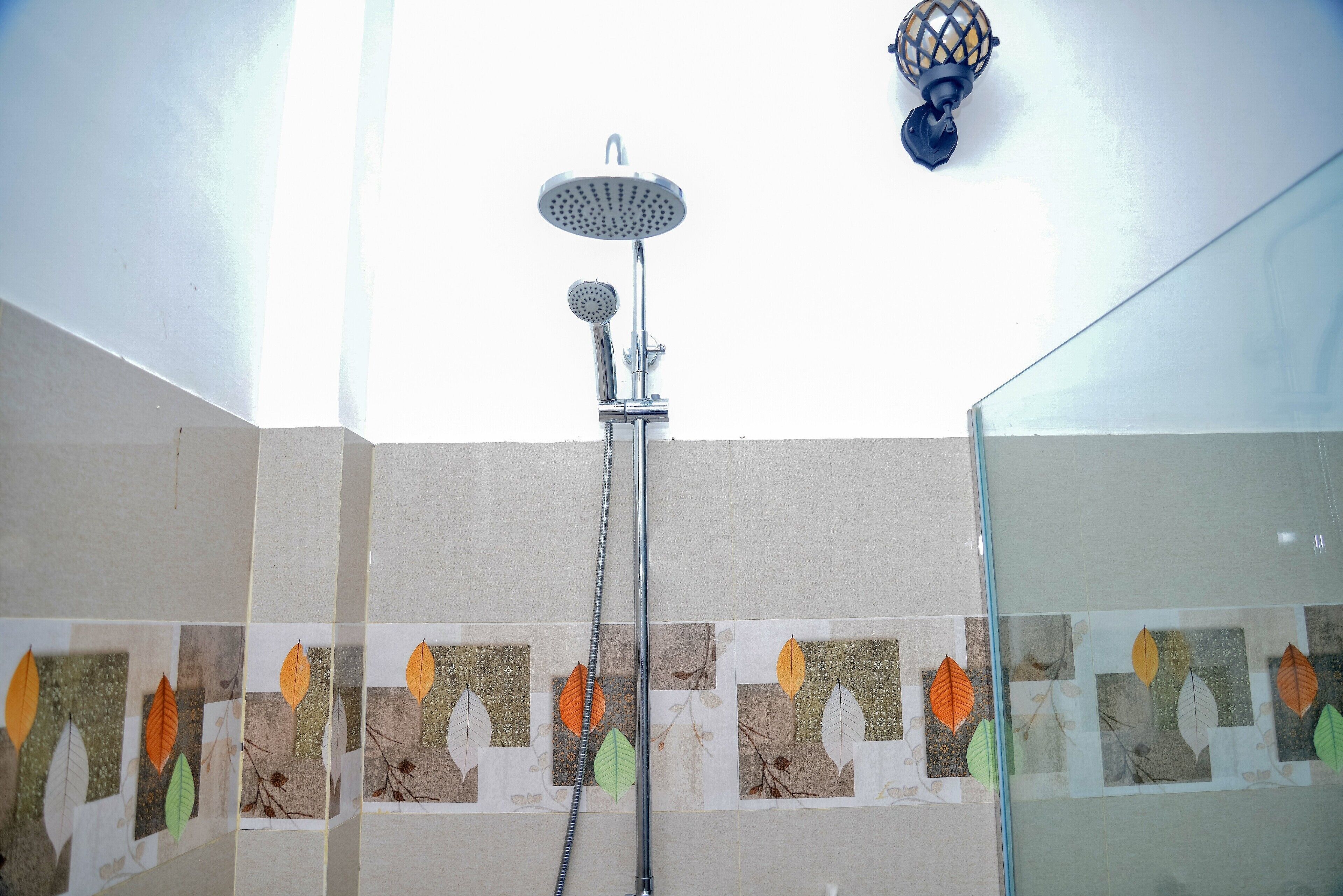 Una ducha con cabezal de ducha de lluvia y cabezal de ducha de mano, cerramiento de ducha de vidrio y una pared con mosaicos con diseño de hojas.