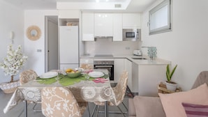 Cocina compacta con armarios blancos, microondas y tostadora. Hay una mesa de comedor con platos, un bol de fruta y una planta en maceta.