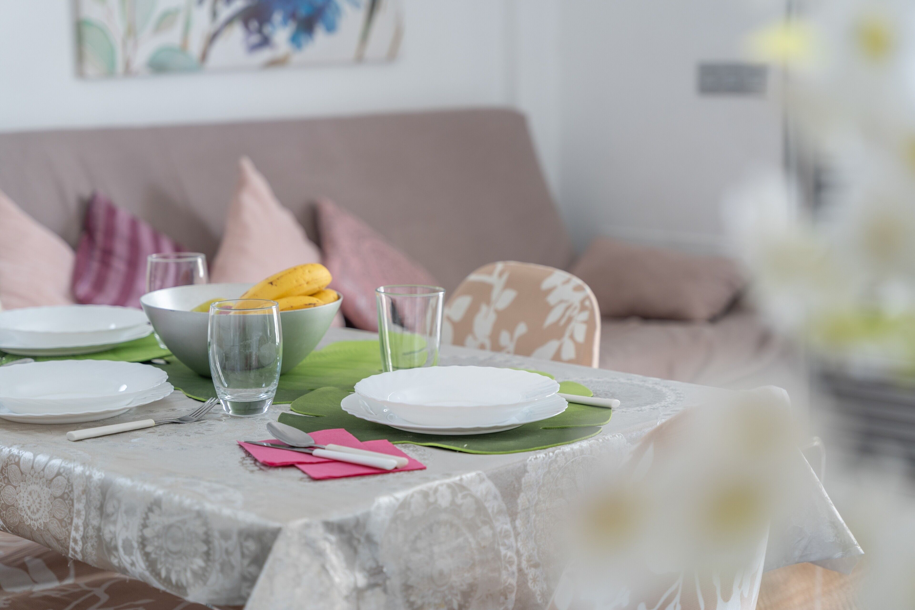 A dining table set with plates, glasses, and cutlery, with a sofa and cushions in the background.