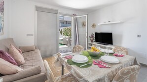 Un área de comedor con una mesa lista para cuatro, un sofá, un televisor y una puerta corredera que da a un espacio exterior.