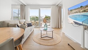 A modern living room with a sofa, dining table, and a TV displaying a pool view. - AL4-2023 ESTEPONA - APARTMENT - ALCAZABA LAGOON (Casares)