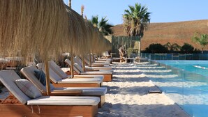 A row of wooden lounge chairs with white cushions, a clear pool, and a sandy beach area. - AL4-2023 ESTEPONA - APARTMENT - ALCAZABA LAGOON (Casares)