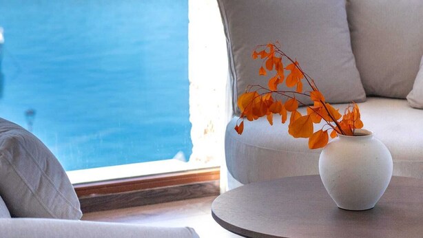 A modern living room with a large window overlooking the ocean, a white sofa, and a round table with a vase of orange leaves.
