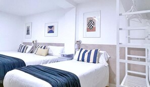 Ein Hotelzimmer mit zwei Betten, jeweils mit einem gestreiften Kopfkissen und einer blauen Decke. An der Wand hängen Bilder in Rahmen und es gibt eine weiße Regaleinrichtung.