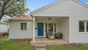 A single-story house with a blue front door, a porch with two wicker chairs and a round table, and a small window on the side.