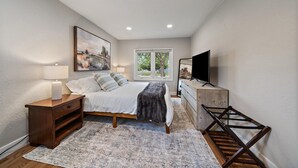 A bedroom with a bed, bedside table, lamp, dresser, TV, and a window with a view of trees.