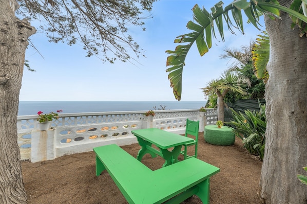 Un patio con mesa y banco, con vista a un cuerpo de agua, rodeado de árboles y plantas.