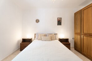 Un dormitorio con una cama grande, mesitas de noche de madera, un armario, un reloj de pared y un cuadro enmarcado.