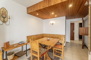 Una habitación de esquina con una mesa de madera, sillas, un banco, una lámpara fijada a la pared y una puerta.