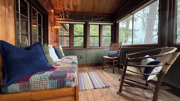 A cozy cabin interior with a wooden log wall, a rocking chair, a cushioned bench with a quilt, and large windows offering a view of the outdoors. - Bay of the Moon at Wolf Point - Pet Friendly! (Crane Lake)