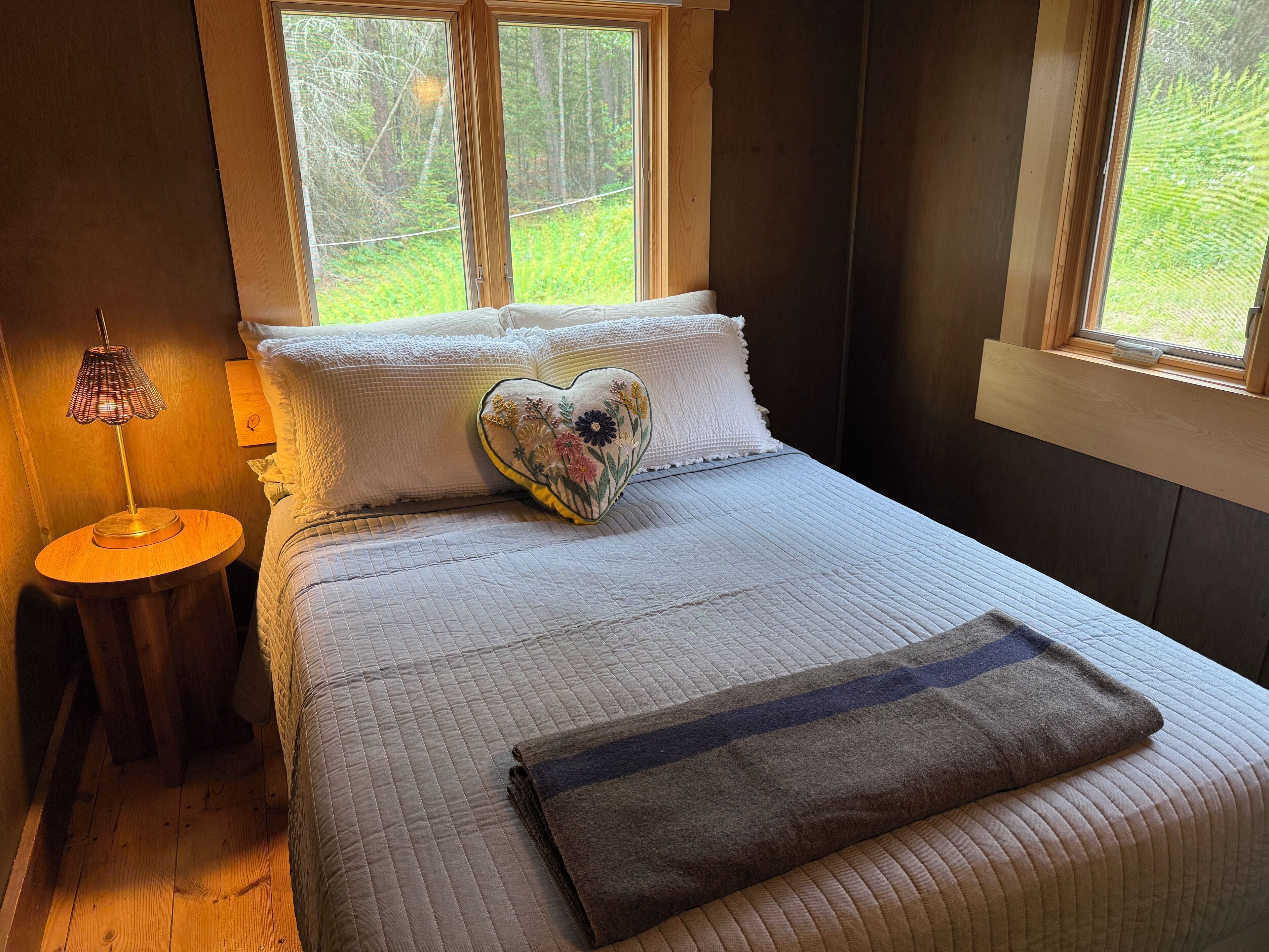 Une chambre à coucher avec un lit, un oreiller en forme de cœur, une table de chevet avec une lampe et des fenêtres donnant sur un paysage verdoyant.