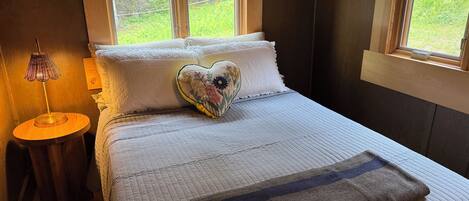 Une chambre à coucher avec un lit, un oreiller en forme de cœur, une table de chevet avec une lampe et des fenêtres donnant sur un paysage verdoyant.