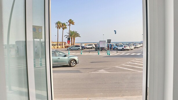 A parking lot with cars and palm trees, a beach in the background, and a clear sky.