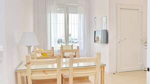 A room with a glass-top dining table, wooden chairs, a lamp, and a TV on the wall.