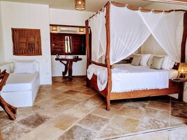 A four-poster bed with a canopy, a wooden nightstand, a small table, and a white sofa in a room with tiled flooring.