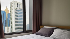 A hotel room with a bed, a window with curtains, and a view of tall buildings.