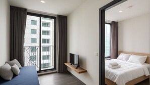 A modern hotel room with a bed, a sofa, a TV on a shelf, and a balcony with curtains.