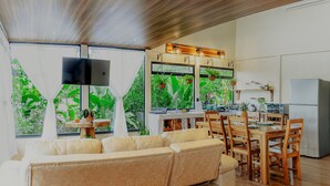 Ein modernes Wohnzimmer mit einer großen beigen Couch, einem Holzespeisetisch und Stühlen. Eine Küchenzeile mit Kühlschrank, Herd und Spüle ist ebenfalls vorhanden. Große Fenster mit Vorhängen bieten Blick ins Grüne.